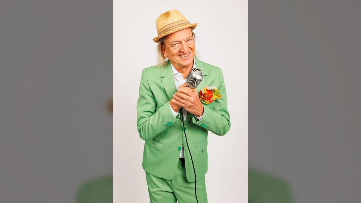 Markus Linder trägt einen hellgrünen Anzug mit Muster, weißes Hemd, Strohhut und ein orange-gelbes Einstecktuch, während er ein Vintage-Mikrofon hält